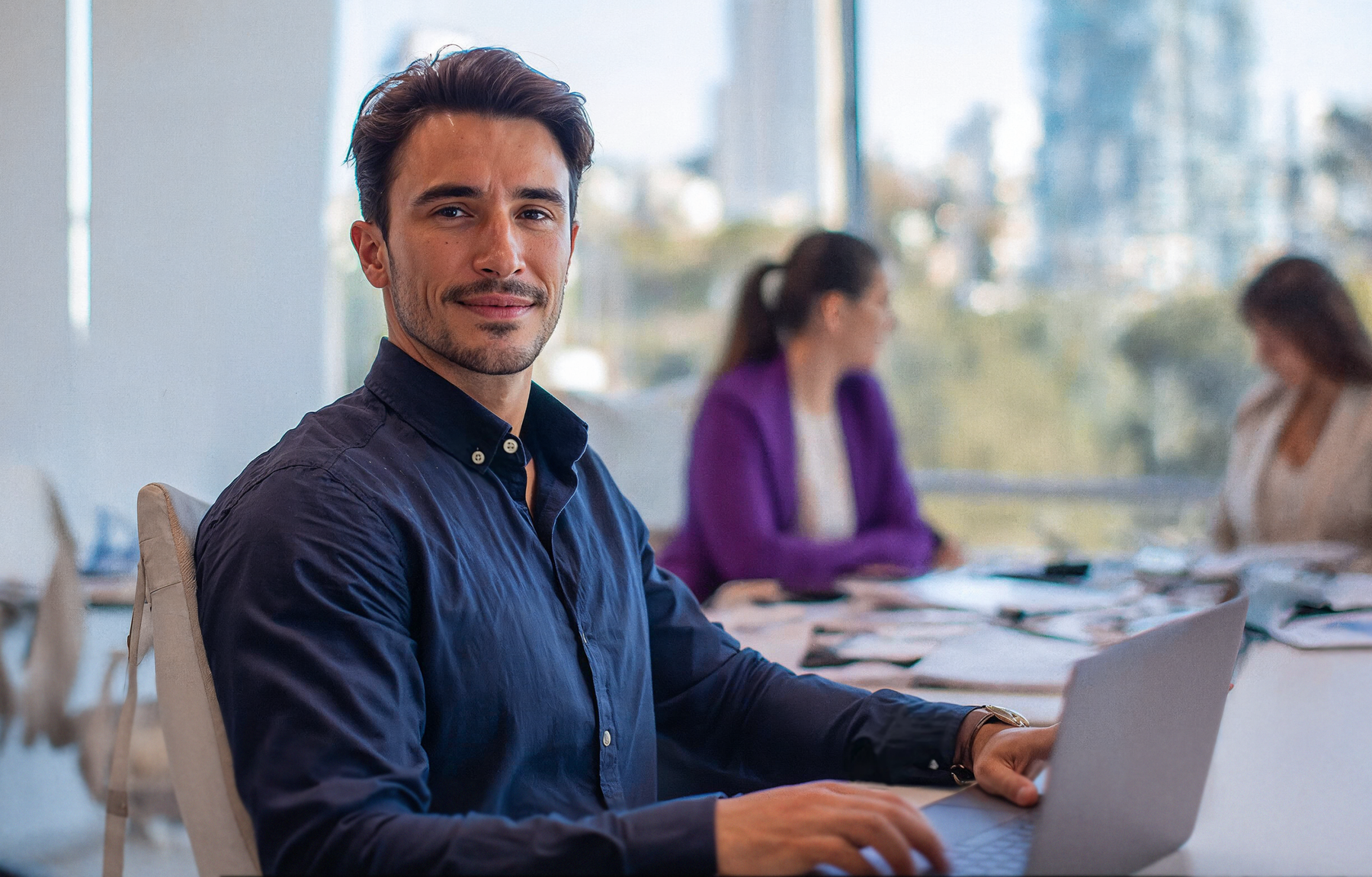 AI-generated business campaign photo: a man in a blue shirt smiles at the camera while working on a laptop at a white table, with two women out of focus in a bright office and city buildings behind the window