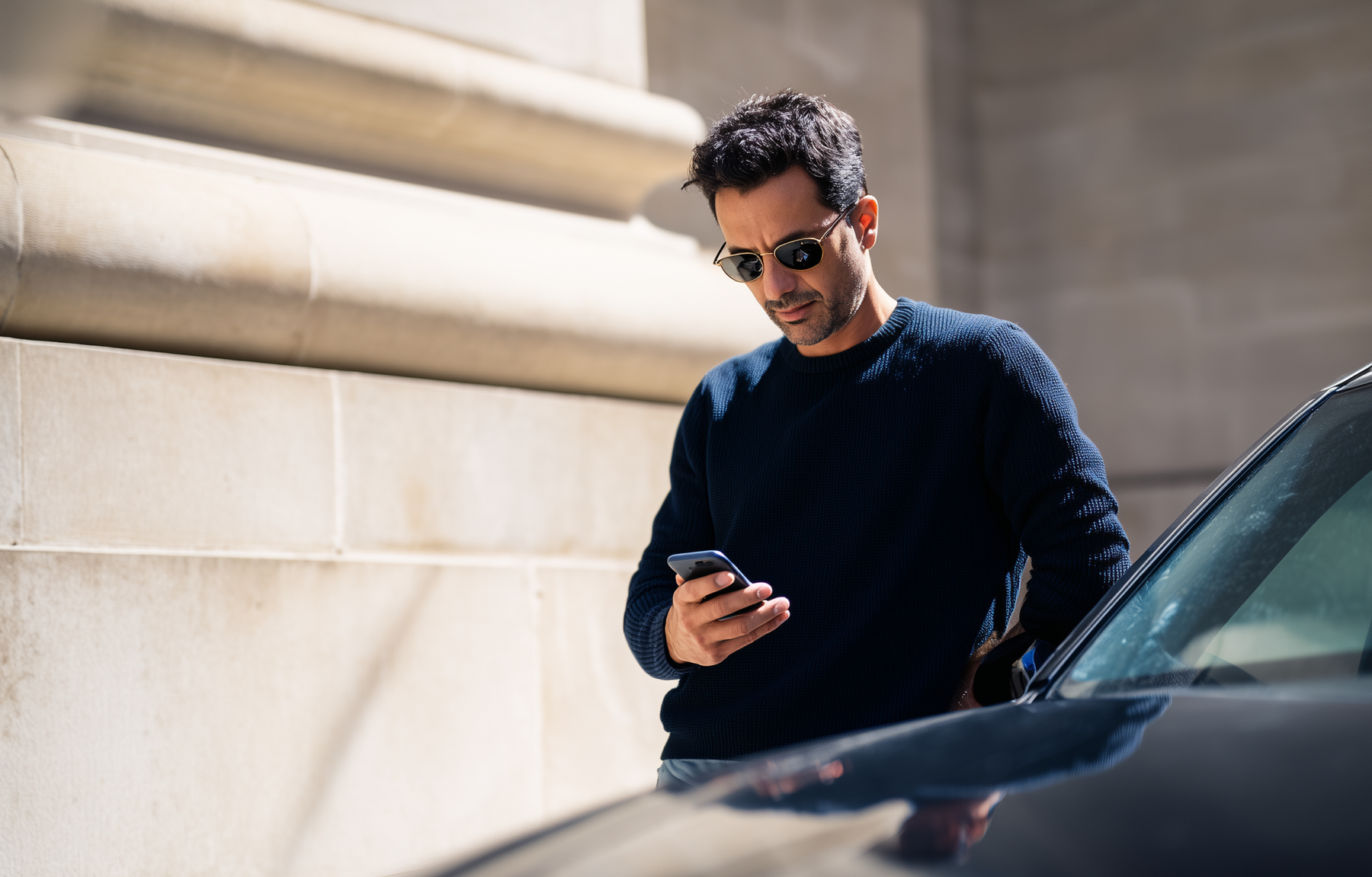 AI-generated driver campaign photo: a man in sunglasses and a dark blue jumper leans on a black car and looks at his phone, in front of a stone building in bright sunlight