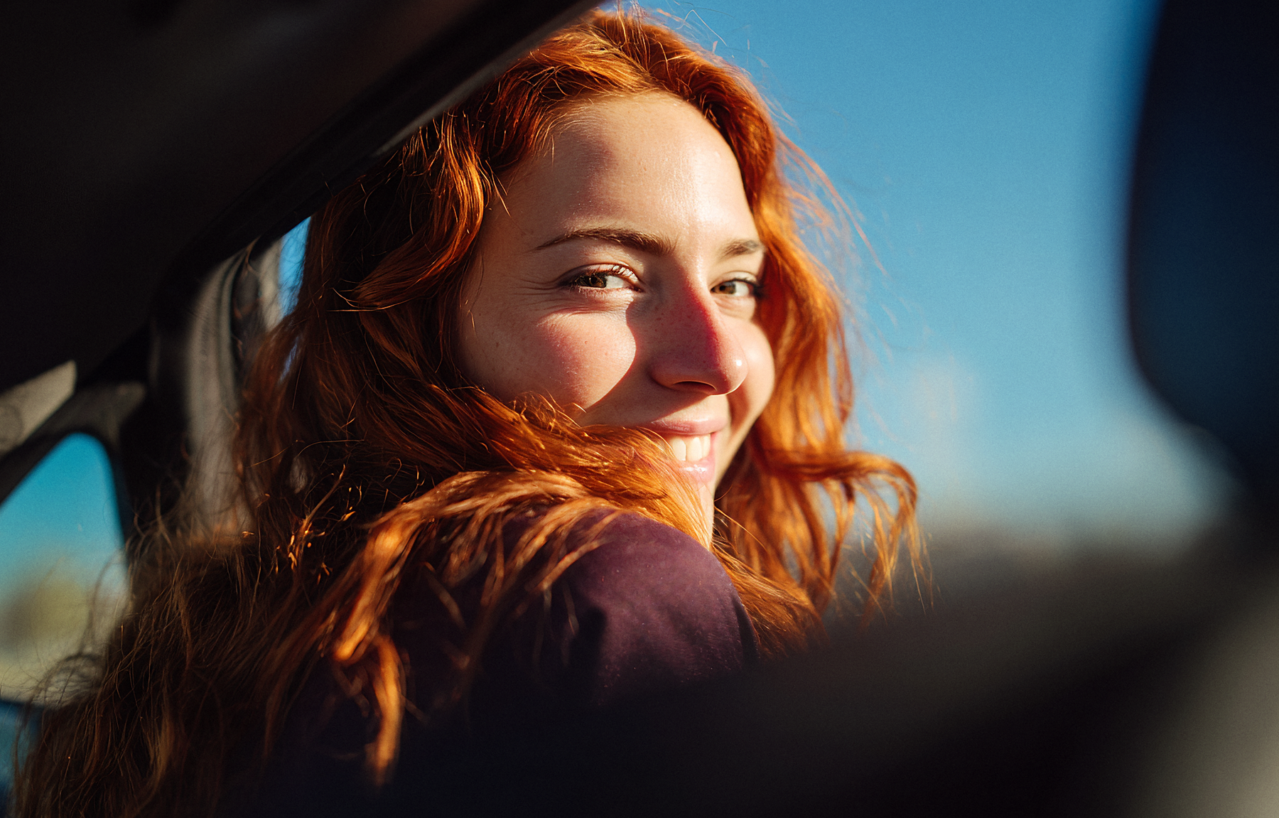 AI-generated rider campaign photo: a smiling woman with long red hair looks over her shoulder at the camera from the back seat of a car, lit by the sun